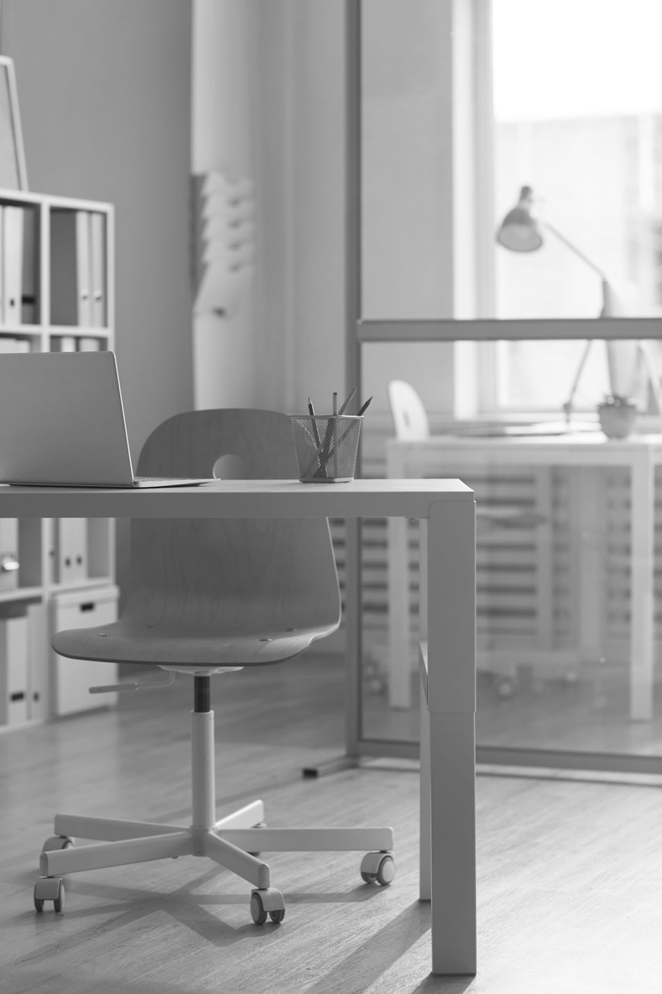 A clean, modern office workspace with a minimalistic design. The scene features a desk with a laptop and a pencil holder, and a swivel chair on wheels. In the background, another desk and chair are visible through a glass partition, emphasizing the tidy and organized environment. The overall atmosphere is bright and conducive to productivity, showcasing the effectiveness of Opticlean's commercial cleaning services.